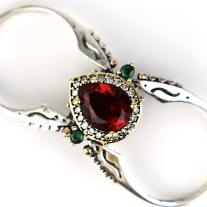 May include: A silver-toned ring with a teardrop-shaped, deep red gemstone at its center. The gemstone is surrounded by a gold setting and smaller white and green stones. The ring has an infinity symbol design.