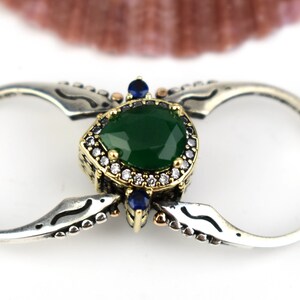 Könnte beinhalten: Ein silberner Doppelring mit einem smaragdgeschliffenen grünen Edelstein in der Mitte, umgeben von kleinen weißen und blauen Steinen. Der Ring hat ein verziertes Design mit dekorativen Details.
