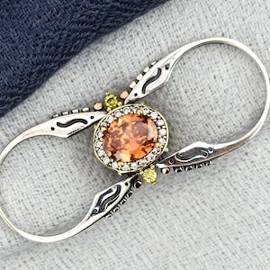Könnte beinhalten: Silberfarbener Ring mit einem ovalen, orangefarbenen Edelstein in der Mitte, umgeben von kleineren weißen und gelben Steinen. Der Ring hat ein offenes Design mit dekorativen Details und liegt auf einem grau-blauen Stoffhintergrund.
