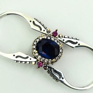 Könnte beinhalten: Ein silberfarbener Ring mit einem ovalen, dunkelblauen Edelstein in der Mitte, umgeben von kleinen weißen Steinen. Der Ring hat dekorative Akzente und zwei Schlaufen auf beiden Seiten. Kleine rote Edelsteine sind ebenfalls vorhanden.