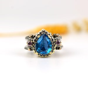 Könnte beinhalten: Ein silberner Ring mit einem großen, birnenförmigen, leuchtend blauen Edelstein als Mittelpunkt. Der Ring hat eine goldfarbene Lünette und ist mit kleineren lila Edelsteinen und goldfarbenen Details verziert. Der Ring ist vor einem weißen Hintergrund platziert.