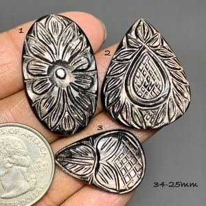 May include: Three dark, polished, carved stone cabochons with intricate floral and geometric patterns. One oval piece, one large teardrop, and one small teardrop are shown next to a US quarter coin for scale, with text indicating 34-25mm sizes.