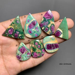 May include: Seven polished cabochons featuring green fuchsite with embedded magenta ruby crystals. The stones are numbered one through seven and vary in shapes including triangles, teardrops, and rectangles. The text 32-24mm is visible.