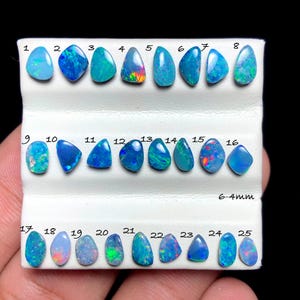 May include: A collection of twenty-five loose, polished opals arranged in three rows on a white display card. Each gemstone is numbered from 1 to 25, displaying various blue, green, and red flashes. The text 6-4mm is printed on the card.