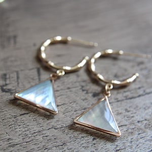 May include: A pair of gold-colored hoop earrings with a textured design. Each earring features a triangular pendant with a pearlescent, iridescent finish and a gold-colored border. The earrings are displayed on a wooden surface.
