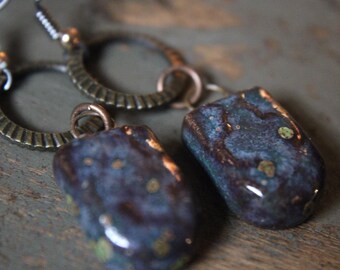 May include: A pair of artisan earrings with dark blue, rectangular pendants. The pendants have a glossy finish with hints of gold and green. The earrings feature bronze-colored, textured hoops and silver ear wires.