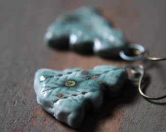 May include: Close-up of two turquoise ceramic earrings. The earrings are leaf-shaped with a textured surface and small gold-colored accents. The earrings are suspended from thin, dark-colored wires.
