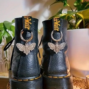 Może przedstawiać: Czarne skórzane buty z srebrnymi zawieszkami w kształcie motyla przywiązanymi do sznurowadeł. Zawieszki są ozdobione misternymi wzorami i mają vintage'owy charakter.