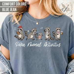 Puede incluir: Camiseta azul vaquero con un diseño de mapache de dibujos animados. El diseño incluye mapaches en varias poses con el texto "Para Normal Activities". La camiseta tiene cuello redondo y mangas cortas.