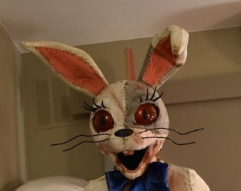May include: A person wearing a white and brown rabbit costume with red eyes and a blue bow tie. The costume is torn and bloody. The person is holding a knife in one hand and a black cord in the other.