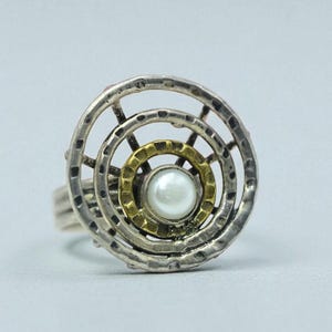 May include: A silver ring with a circular design. The ring features concentric circles, with a gold-colored center and a pearl at the core. The outer circles have a textured, hammered appearance.