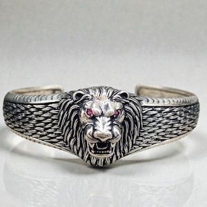 May include: A silver-toned cuff bracelet featuring a detailed lion's head with red gemstone eyes. The lion's mane is intricately designed, and the bracelet has a textured band. A bold piece of jewelry.