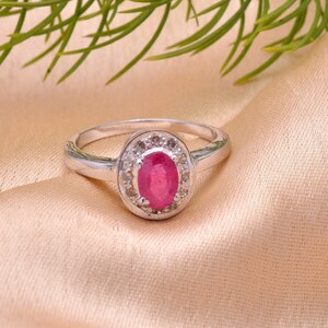 May include: A silver ring featuring an oval ruby gemstone at its centre, encircled by smaller clear stones. The ring rests on peach-coloured satin, with green foliage in the background. A classic piece of jewellery.