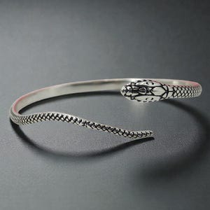May include: A silver-toned cuff bracelet in the shape of a snake. The snake's head is detailed with scales and eyes, while the body tapers to an open end. The bracelet is designed to wrap around the wrist, offering a unique and stylish accessory.
