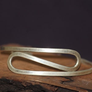 Könnte beinhalten: Ein silberfarbenes Metallarmband mit minimalistischem, offenem Design. Das Armband hat eine durchgehende, fließende Form mit abgerundeten Enden. Es liegt auf einer dunklen Holzoberfläche mit dunklem Hintergrund.