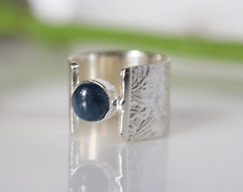 May include: A silver ring with a blue gemstone. The ring has a wide band with an etched floral pattern on the sides. The blue gemstone is round and set in a silver bezel.
