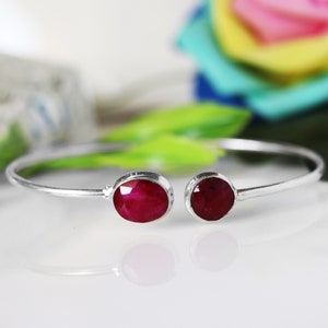Könnte beinhalten: Ein silbernes Armreif-Armband mit zwei roten Edelstein-Akzenten. Das Armband ist offen und hat ein schlichtes Design.