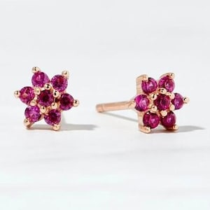 May include: A pair of flower-shaped stud earrings. Each earring has a cluster of small, round, magenta gemstones set in a gold-coloured metal. The earrings are displayed on a white surface.