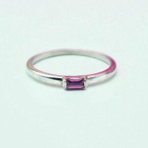 Peut inclure: Une bague en argent avec une pierre précieuse rectangulaire rose foncé. Le bracelet est fin et délicat, et la pierre est sertie dans un style simple et moderne. La bague est un bijou.