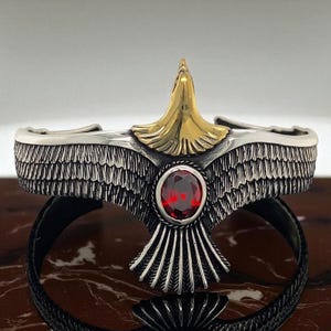 May include: Silver cuff bracelet with an eagle design, featuring a gold head and a red gemstone. The eagle's wings are detailed with feather patterns. The bracelet has a black band.