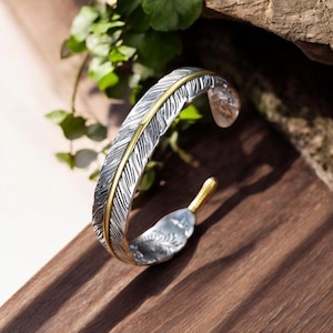 May include: A silver-coloured cuff bracelet with a feather design. The bracelet has a gold-coloured accent down the centre of the feather and at the tip. The bracelet is displayed on a wooden surface with greenery in the background.