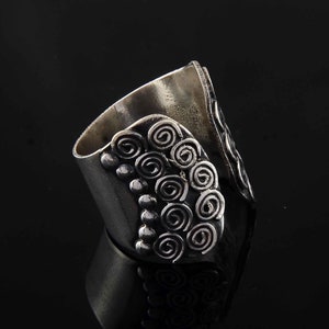 Könnte beinhalten: Ein silberfarbener Ring mit breitem Band und offenem Design. Der Ring hat Spiral- und Punktmuster auf der Vorderseite und eine glatte, gebogene Oberfläche auf der Rückseite. Der Ring wird vor schwarzem Hintergrund gezeigt.