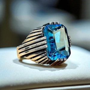 May include: A gold and silver ring featuring a large, rectangular, light blue gemstone. The ring has a detailed design with a textured band and a decorative setting around the gemstone. The ring is set against a neutral background.