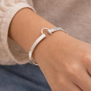 May include: A silver-toned metal cuff bracelet featuring a textured, hammered surface finish. The bracelet has a C-shaped design with a small, hinged mechanical clasp mechanism at the top, allowing the band to open and close securely on the wrist.