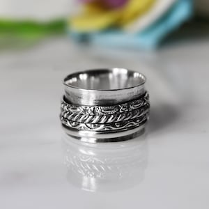 May include: A silver-colored spinner ring with a textured design. The ring features multiple bands, with the central band displaying an intricate floral pattern. The ring's design includes a spinning top section, adding a unique element to the jewelry.