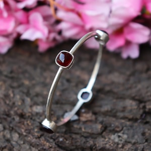 Könnte beinhalten: Ein silbernes Armreif-Armband mit drei roten Edelstein-Akzenten. Das Armband ist offen und hat ein schlichtes Design.