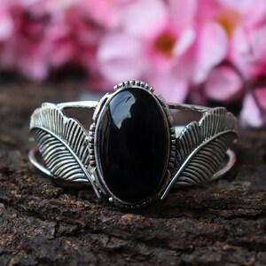 May include: Silver-toned cuff bracelet featuring a large, oval black gemstone. The bracelet has a detailed feather design on each side of the gemstone, set against a dark wood background. The gemstone is approximately 1.5 inches long.