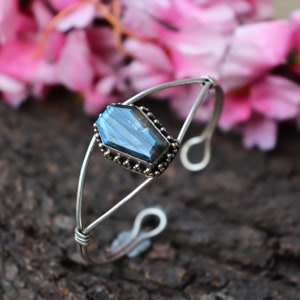 Könnte beinhalten: Ein silberfarbenes Armreif-Armband mit einem rechteckigen, facettierten blauen Edelstein. Der Edelstein ist in einer Perlenfassung gefasst. Das Armband hat ein schlichtes, offenes Design mit gebogenen Armen und einem Hakenverschluss. Rosa Blumen im Hintergrund.