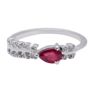 Könnte beinhalten: Silberring mit einem tropfenförmigen, tiefroten Edelstein als Mittelpunkt. Das Ringband weist ein blattartiges Design mit kleinen, klaren Akzentsteinen auf. Der Ring ist vor einem weißen Hintergrund platziert.