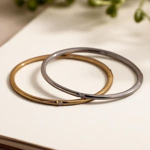 Könnte beinhalten: Zwei elegante Armreifen, einer goldfarben und der andere silberfarben, jeweils mit einem kleinen, funkelnden Akzent. Die Armbänder sind vor einem neutralen Hintergrund platziert, was ihr schlichtes, aber raffiniertes Design hervorhebt. Ein perfektes Accessoire für den Alltag.