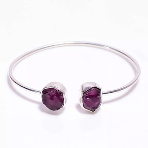 May include: A silver cuff bracelet with two rough-cut, deep purple gemstones. The gemstones are set in silver bezels at the open ends of the bracelet. The bracelet is shown against a white backdrop.