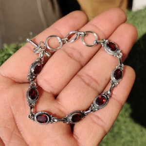 May include: A silver bracelet with a chain link design and red oval-shaped gemstones. The bracelet has a lobster claw clasp.