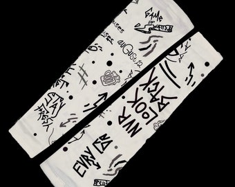 Pode incluir: Um par de aquecedores de braço brancos com desenhos pretos em estilo graffiti. Os desenhos incluem palavras como "Stay Fresh", "Every Day", "Never Look Back" e "Game On".