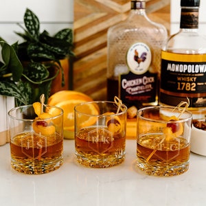 May include: Three personalized old fashioned glasses with a hexagonal design and initials. The glasses are filled with a brown liquid and garnished with orange slices and cherries. Two bottles of whiskey are in the background.