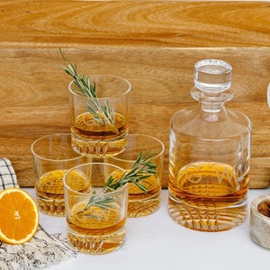 May include: A set of four clear glass whiskey glasses with a decorative cut design, a clear glass decanter with a stopper, and a wooden serving tray with a cut-out handle. The glasses and decanter are filled with amber liquid. There are two orange slices and a small bowl of nuts on the tray.