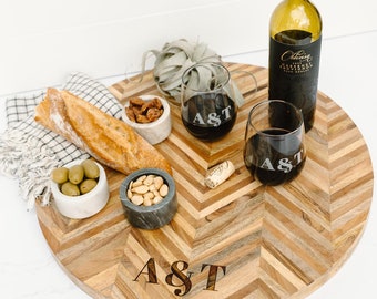 May include: A round wooden serving board with a herringbone pattern, holding wine glasses, a bottle of wine, and small bowls of snacks. The wine glasses are engraved with "A & T". The board is approximately 20 inches in diameter.