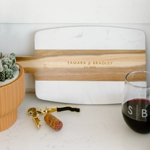 May include: A white and brown marble cutting board with the text "Samara & Bradley est. 2023" engraved on the wood. Two wine glasses with the initials "SB" etched on them, a bottle of Cabernet-Syrah wine, a corkscrew, and a cork are on a white surface.
