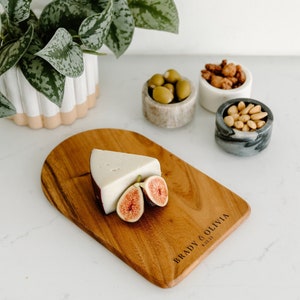 May include: A wooden serving board with a piece of cheese and sliced figs. The board is engraved with the names "BRADY & OLIVIA" and the date "6.22.23". Small marble bowls hold olives, nuts, and candied nuts. A potted plant is in the background.
