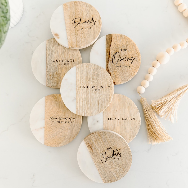 Puede incluir: Conjunto de ocho posavasos redondos de madera con parte superior de m&aacute;rmol blanco. Cada posavasos tiene una inscripci&oacute;n personalizada con un nombre y una fecha. Los posavasos est&aacute;n dispuestos en c&iacute;rculo sobre una superficie blanca.