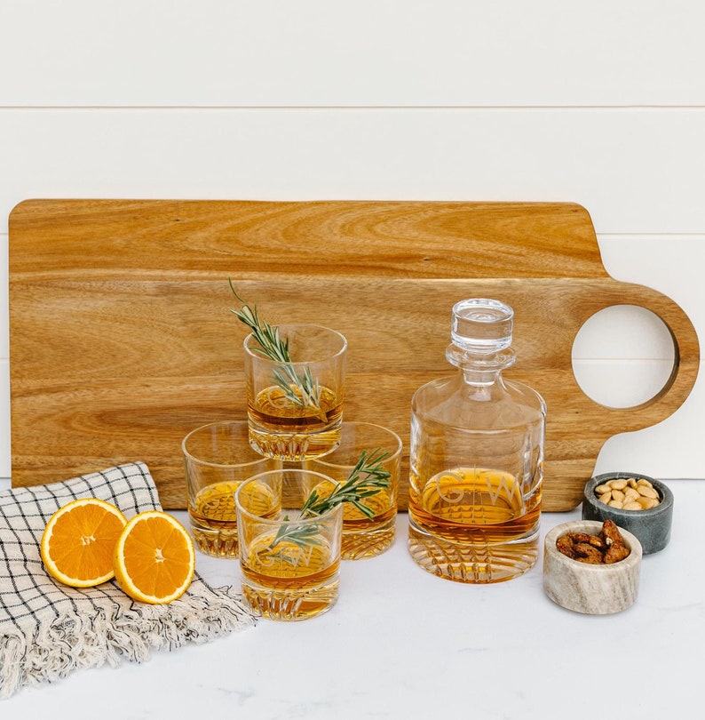 May include: A set of four clear glass whiskey glasses with a decorative cut design, a clear glass decanter with a stopper, and a wooden serving tray with a cut-out handle. The glasses and decanter are filled with amber liquid. There are two orange slices and a small bowl of nuts on the tray.