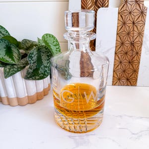 May include: A clear glass decanter with a stopper and a diamond-shaped pattern. The decanter is filled with a golden liquid and has the initials "G.W." etched on the front.
