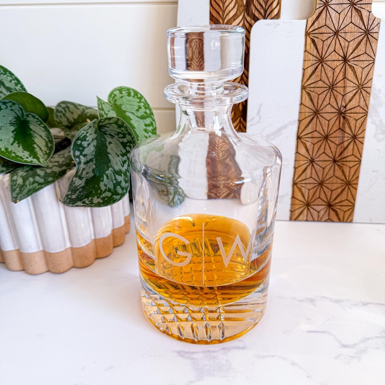 May include: A clear glass decanter with a stopper and a diamond-shaped pattern. The decanter is filled with a golden liquid and has the initials "G.W." etched on the front.