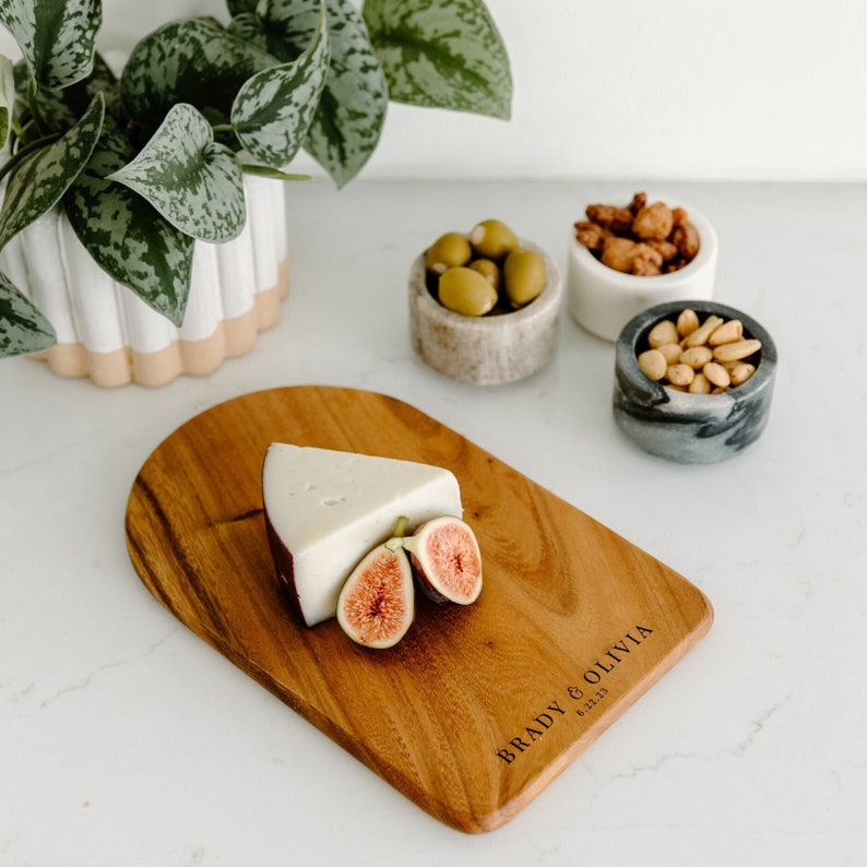 May include: A wooden serving board with a piece of cheese and sliced figs. The board is engraved with the names "BRADY & OLIVIA" and the date "6.22.23". Small marble bowls hold olives, nuts, and candied nuts. A potted plant is in the background.