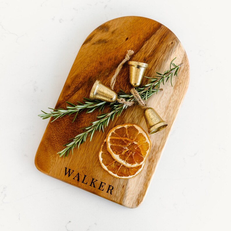 May include: A wooden serving board with rounded edges, engraved with the name "WALKER". It is decorated with three small gold bells, sprigs of rosemary, and two dried orange slices. The board is made of a light and dark brown wood.