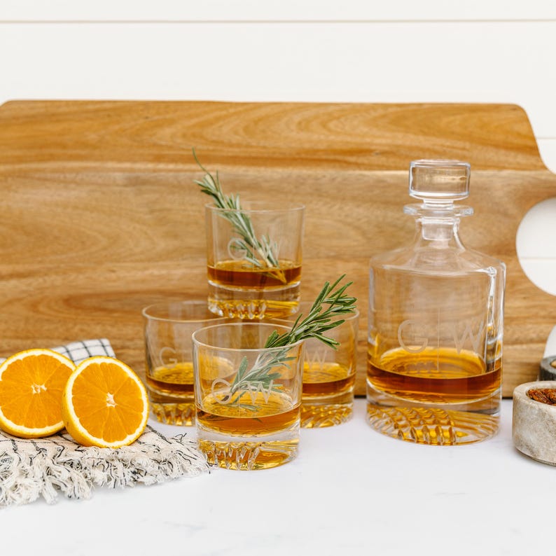 May include: Set of four clear glass whiskey tumblers with a decorative etched design and a decanter filled with amber liquid. The glasses and decanter are on a wooden cutting board with orange slices and sprigs of rosemary.