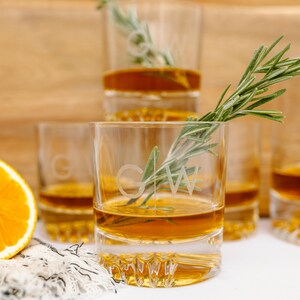 May include: Four clear glass tumblers with a textured bottom, filled with a golden liquid. The tumblers are engraved with the initials "G.W." and a sprig of rosemary is placed in the front tumbler.